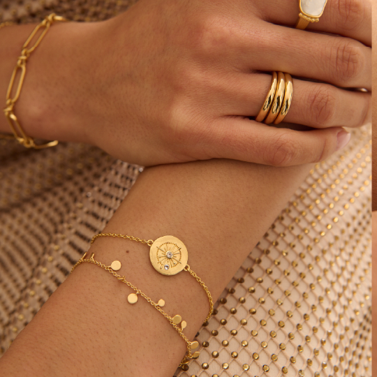 Close-up of a hand wearing gold jewelry including bracelets and rings on a textured beige background.