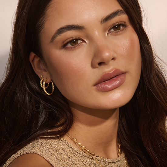 Close-up of a woman wearing gold hoop earrings and a gold necklace against a neutral background