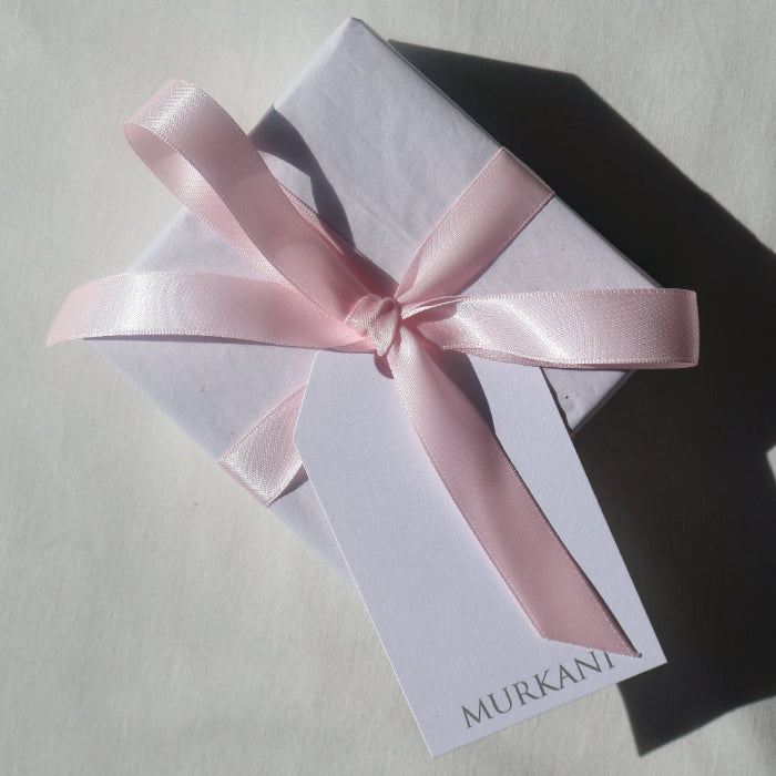 A white rectangular box tied with a light pink satin ribbon bow, with a white card hanging from the front that reads MURKANI, photographed on a light neutral surface with strong sunlight and shadow.
