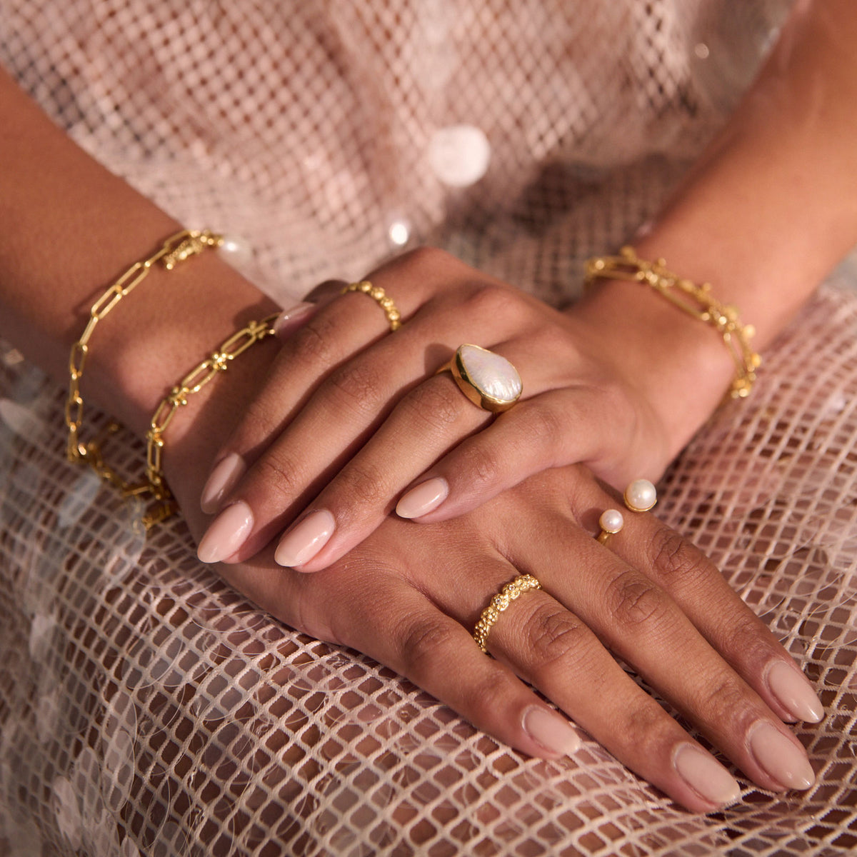 A close-up of crossed hands wearing multiple gold rings and bracelets, including a ring with a large white stone and pearl accents, set against a shimmering pink mesh fabric background.
