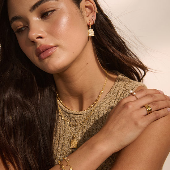 Woman wearing gold jewelry including earrings, necklace, ring, and bracelet against a neutral background.